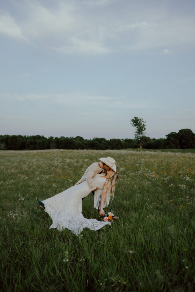 bride and groom share dip kiss in field of wildflowers at rustic oklahoma wedding venue