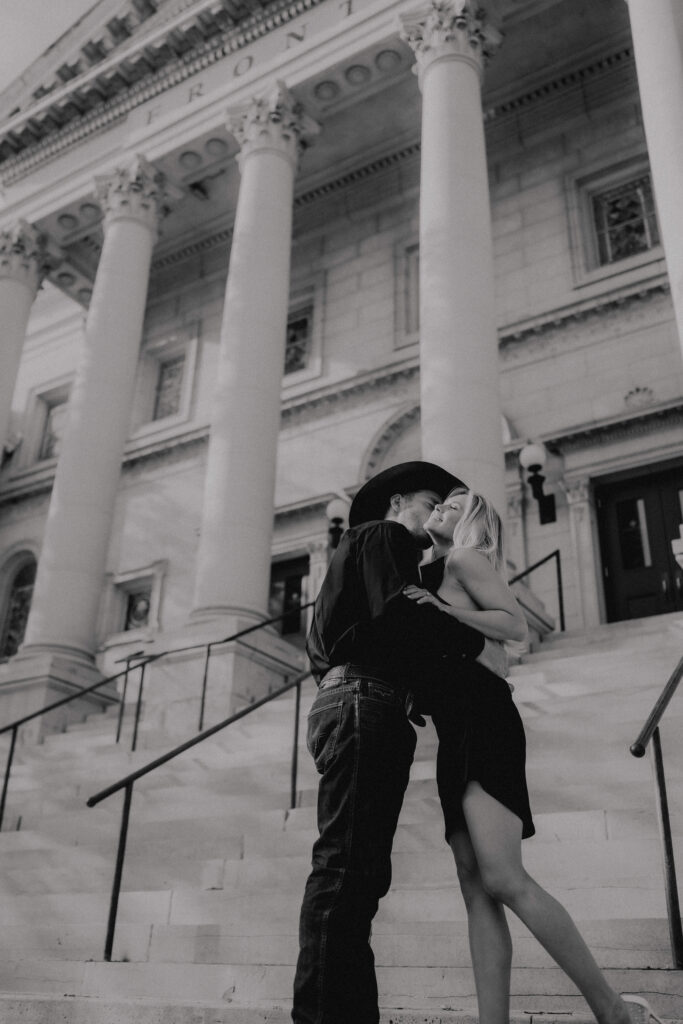 he kisses her on the cheek as she smiles out front of a building in downtown okc