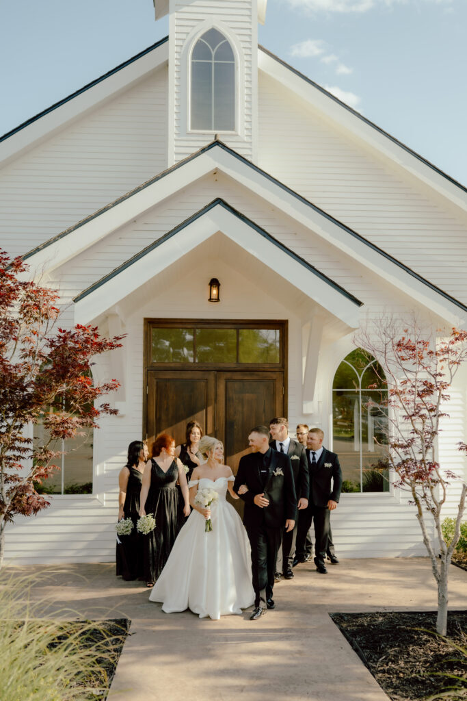 bride and groom walk with their wedding party outside white chapel wedding venue