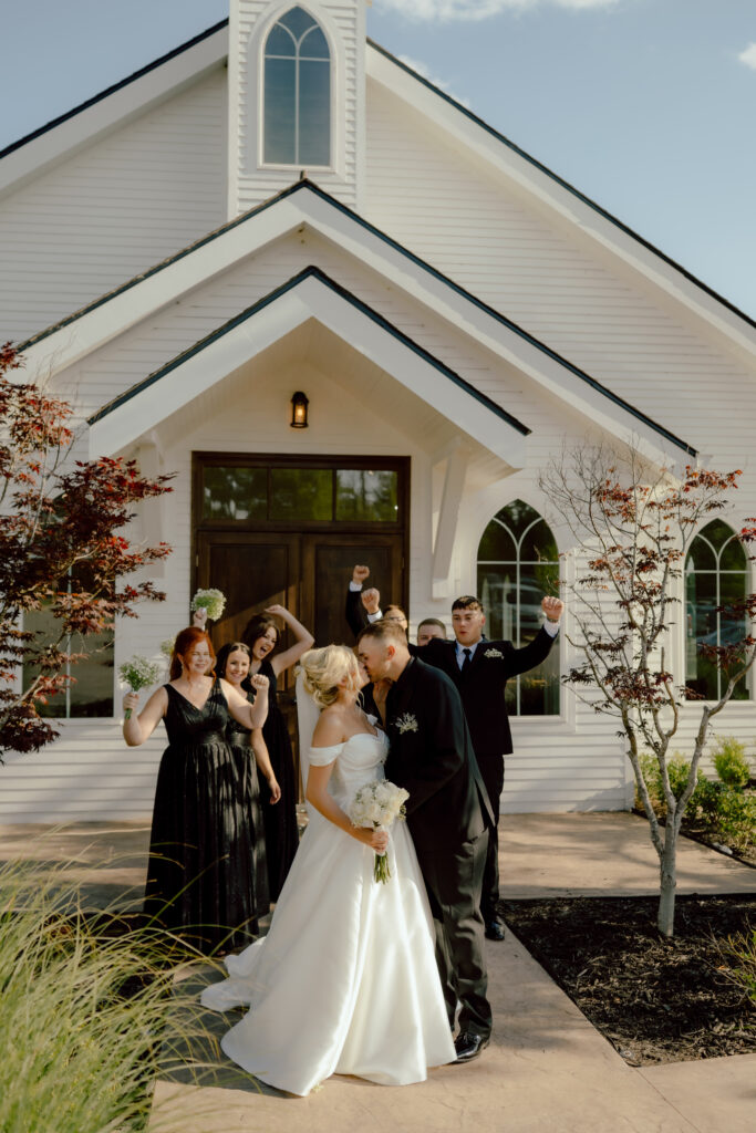 bride and groom kiss as wedding party cheers outside white chapel